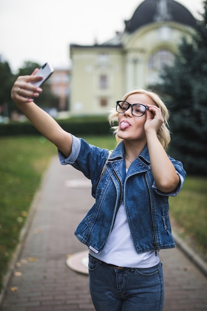 Mulher loira de moda elegante em jeans e óculos fazendo selfie em seu telefone na cidade pela manhã
