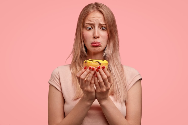 Foto grátis mulher loira descontente segura uma rosquinha doce, parece tentada, quer comer, mas continua fazendo dieta, vestida com uma camiseta casual, isolada sobre a parede rosa. pessoas e conceito de junk food.