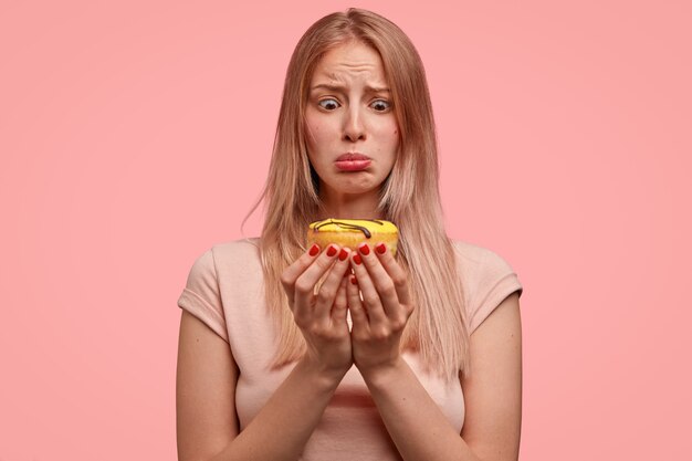 Mulher loira descontente segura uma rosquinha doce, parece tentada, quer comer, mas continua fazendo dieta, vestida com uma camiseta casual, isolada sobre a parede rosa. Pessoas e conceito de junk food.