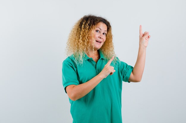 Mulher loira e bonita apontando para cima em uma camiseta polo verde e parecendo surpresa, vista frontal.
