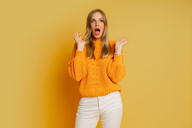 Mulher loira e bonita com rosto suprice em suéter laranja elegante de outono posando em amarelo.