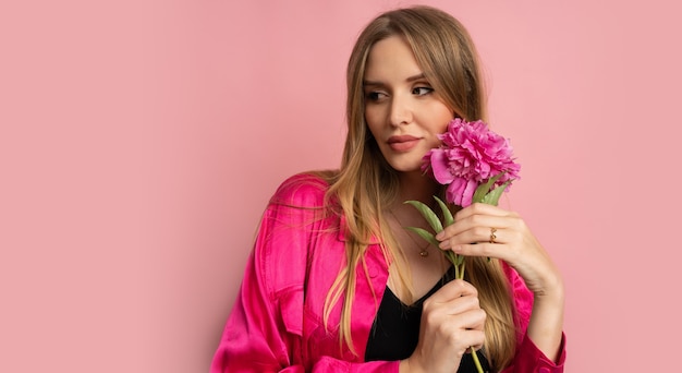 Mulher loira e bonita posando com uma flor de peônia em uma roupa elegante de verão sobre uma parede rosa