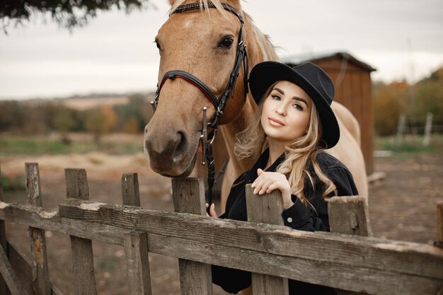 Mulher loira e cavalo marrom em uma fazenda. Mulher vestindo roupas pretas e chapéu. Mulher tocando o cavalo atrás da cerca.