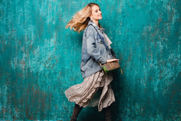 Mulher loira elegante atraente e sexy em jeans e jaqueta grande caminhando contra uma parede verde vintage na rua