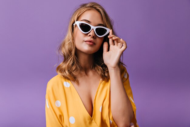 Mulher loira elegante com roupas amarelas, posando com elegantes óculos de sol. Modelo feminino caucasiano sério em pé roxo.