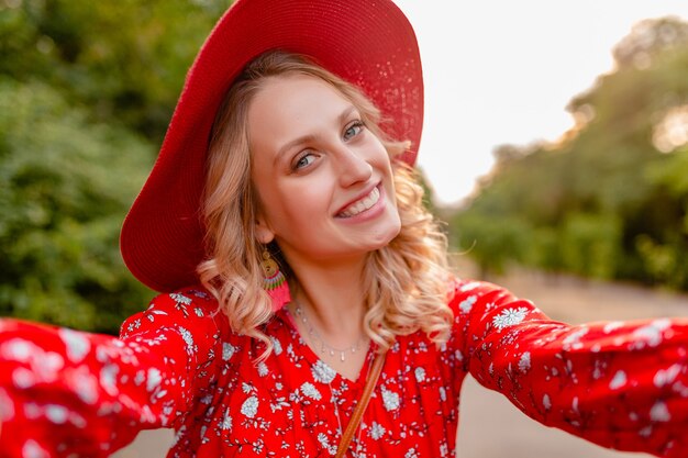 Mulher loira elegante e atraente e sorridente com chapéu vermelho palha e blusa, roupa da moda de verão, tirando foto de selfie