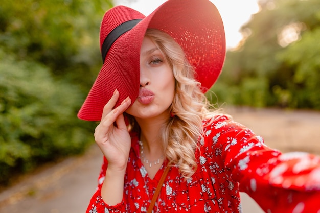 Mulher loira elegante e atraente e sorridente com chapéu vermelho palha e blusa, roupa da moda de verão, tirando foto de selfie