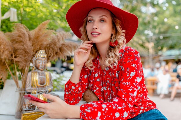Mulher loira elegante e atraente sorridente com chapéu vermelho palha e blusa, roupa da moda de verão segurando usando smart phone cafe