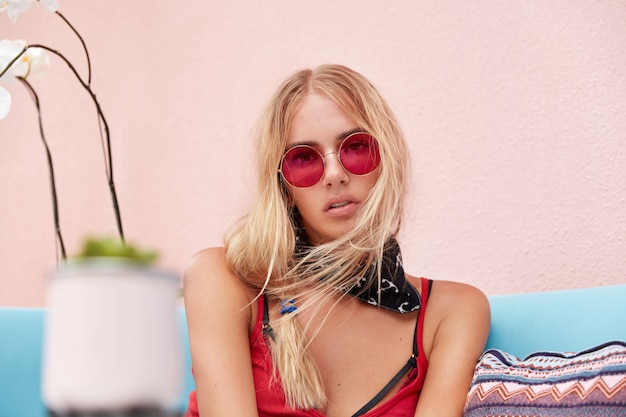 mulher loira em óculos de sol da moda, usa roupas da moda e óculos de sol vermelhos, senta-se contra a parede rosa em um sofá confortável.