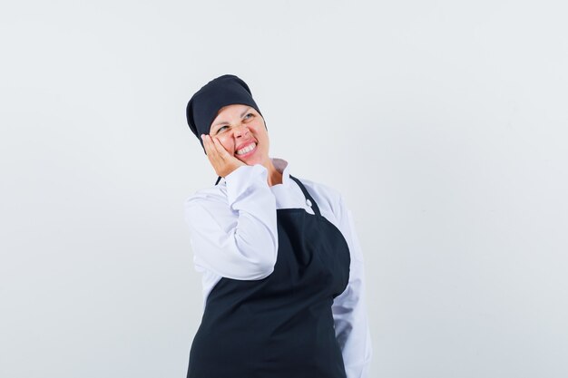 Mulher loira em pose de pensamento, inclinando a bochecha por lado em uniforme preto de cozinheiro e olhando pensativa.