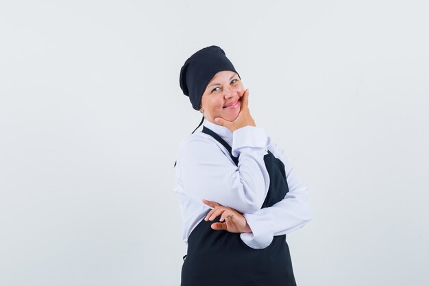 Mulher loira em pose pensativa, colocando a mão na bochecha com uniforme preto de cozinheira e bonita