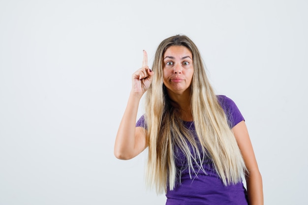 mulher loira em t-shirt violeta, apontando para cima e olhando curiosa, vista frontal.