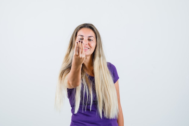 mulher loira em t-shirt violeta, fazendo gesto italiano e olhando alegre, vista frontal.