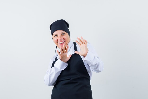 Mulher loira em uniforme de cozinheiro preto, esticando as mãos como algo parando e olhando bonita, vista frontal.