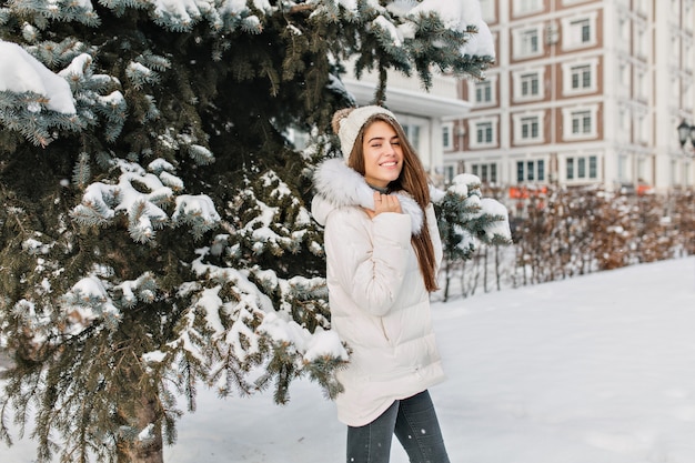 Mulher loira encantadora com jaqueta branca e calça jeans preta, posando durante um passeio em winter park. Foto ao ar livre de uma mulher muito elegante se divertindo na manhã de dezembro.