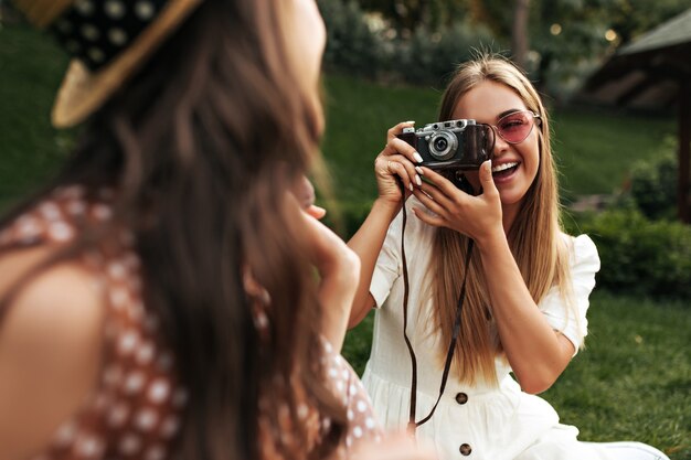 Mulher loira encantadora com vestido branco estiloso e óculos de sol vermelhos, sorri e tira uma foto da amiga