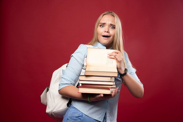 Mulher loira estudante segurando uma pilha pesada de livros e parece cansada.