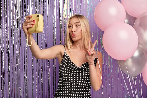 Mulher loira feliz faz maquiagem, mantém os lábios dobrados, faz selfie com pequena câmera, encosta na parede decorada com balões e enfeites, usa vestido estiloso, mostra gesto de paz com a mão.