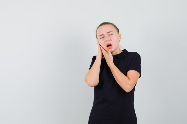 Mulher loira fingindo estar com dor de dente usando uma camiseta preta e parecendo exausta