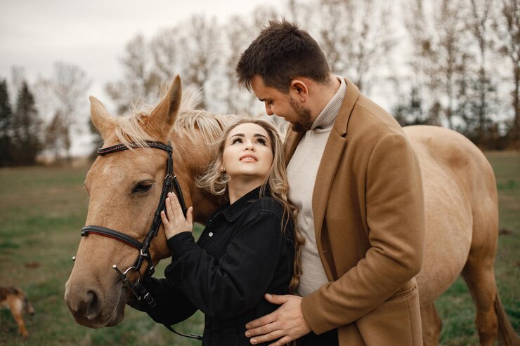 Foto grátis mulher loira, homem moreno de pé no campo com cavalo marrom. mulher vestindo roupas pretas e homem vestindo casaco marrom. homem e mulher abraçados.