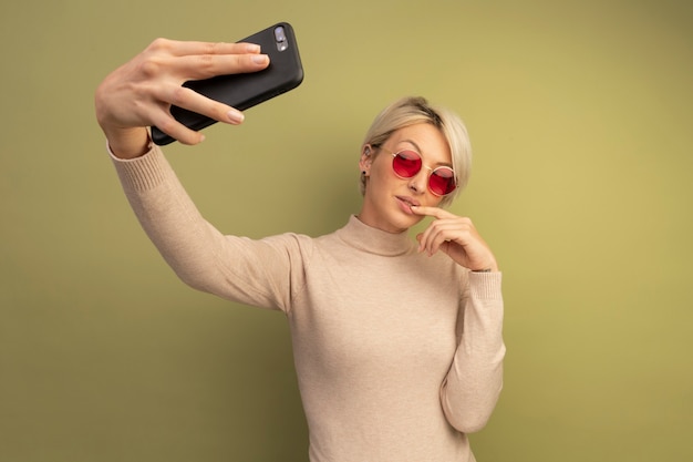 Mulher loira jovem confiante usando óculos escuros, colocando o dedo no lábio, tirando uma selfie isolada na parede verde oliva com espaço de cópia