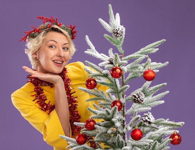 Mulher loira jovem e alegre usando coroa de flores de Natal e guirlanda de ouropel em volta do pescoço, atrás de uma árvore de Natal decorada, olhando para manter a mão sob a cabeça isolada na parede roxa