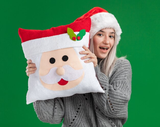 Mulher loira jovem feliz com um suéter de inverno e um chapéu de Papai Noel segurando uma almofada de Natal, olhando para a câmera com um sorriso no rosto, de pé sobre um fundo verde