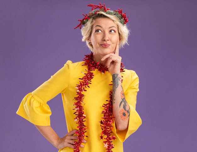 Mulher loira jovem pensativa usando coroa de flores de Natal e guirlanda de ouropel em volta do pescoço, mantendo a mão na cintura e no queixo, olhando para cima isolada no fundo roxo com espaço de cópia