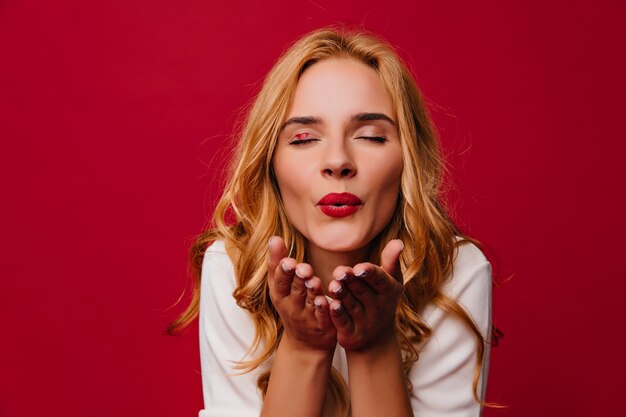 mulher loira maravilhosa mandando beijo no ar. Adorável garota adorável posando na parede vermelha.