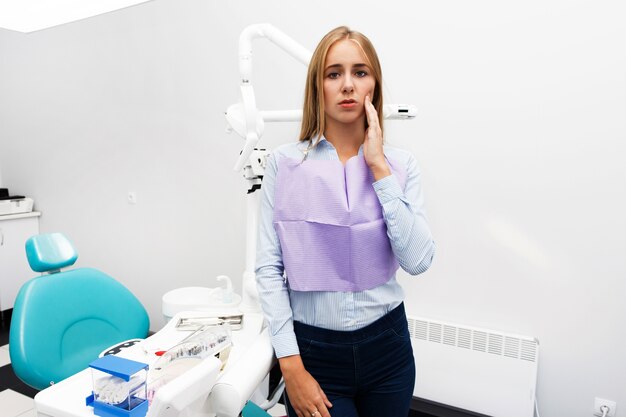 Mulher loira mostra dor de dente em pé no escritório do dentista