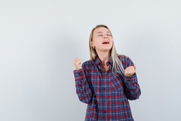 Mulher loira mostrando gesto de sucesso em camisa e olhando feliz, vista frontal.