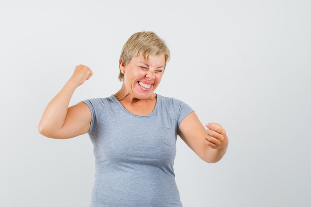 Mulher loira mostrando gesto de vencedor em t-shirt azul claro e olhando alegre, vista frontal.