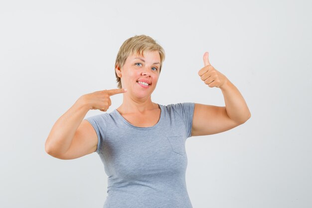 Mulher loira mostrando os polegares e apontando para a mão dela em uma camiseta azul clara e olhando alegre, vista frontal.