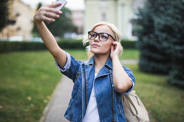 Mulher loira muito elegante e elegante em jeans fazendo selfie em seu telefone na cidade pela manhã