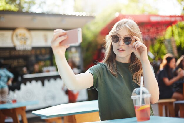 Mulher loira sardenta fazendo uma selfie em copos para postar em suas redes sociais em um dia de verão no parque.