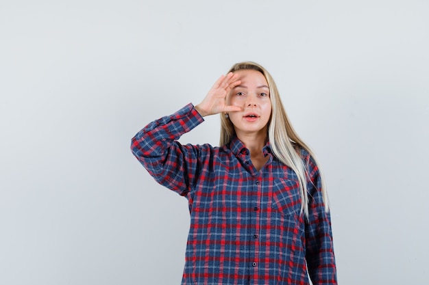 Mulher loira segurando a mão perto do olho em camisa e parece atraente. vista frontal.