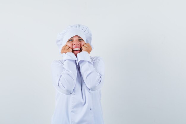 Mulher loira segurando as mãos no queixo, cerrando os punhos em uniforme de cozinheiro branco e está linda.