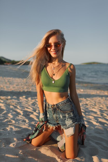 Mulher loira sexy top verde colheita e jeans posando na praia tropical.