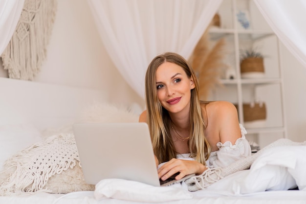 Mulher loira sorridente deitada em uma cama aconchegante e usando o laptop para fazer compras online.