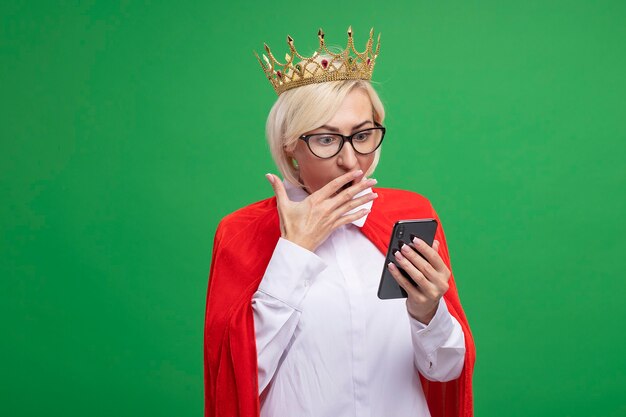 Mulher loira super-heroína de meia-idade preocupada com capa vermelha usando óculos e coroa segurando e olhando para o telefone celular, mantendo a mão na boca isolada na parede verde com espaço de cópia