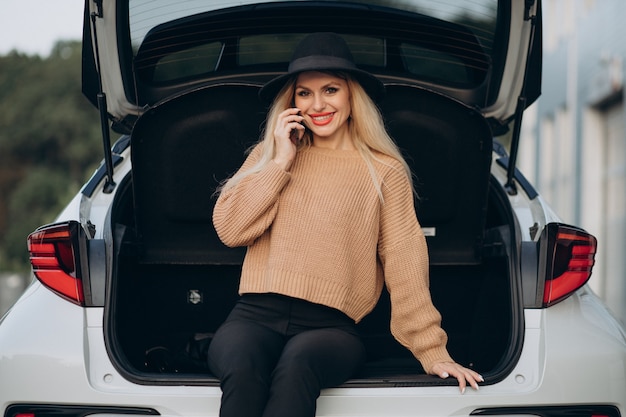 Mulher loira usando o telefone e sentada no porta-malas do carro