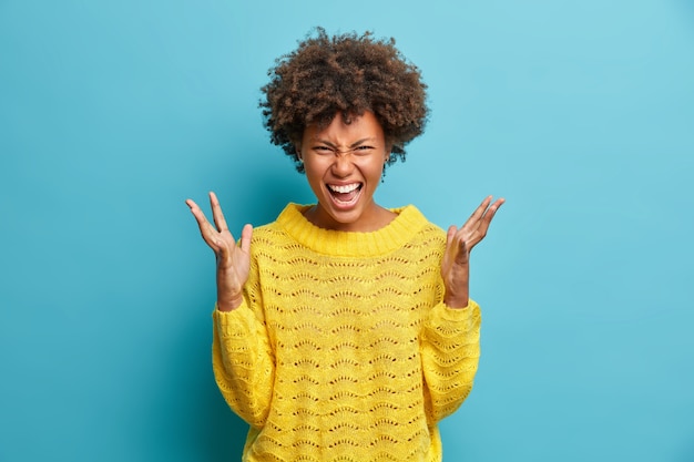 Foto grátis mulher louca e indignada de cabelo encaracolado grita alto e gesticula com raiva, grita furiosamente vestida em poses de suéter de malha amarela contra a parede azul