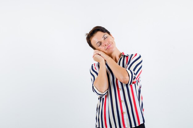 Mulher madura, apoiando a bochecha nas palmas das mãos como travesseiro na camisa listrada e parecendo com sono, vista frontal.