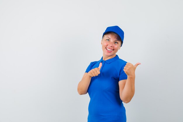 Mulher madura apontando de lado com polegares para cima em t-shirt azul e olhando alegre.