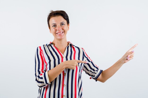 Mulher madura apontando para baixo com o dedo indicador em uma camisa listrada e olhando feliz, vista frontal.