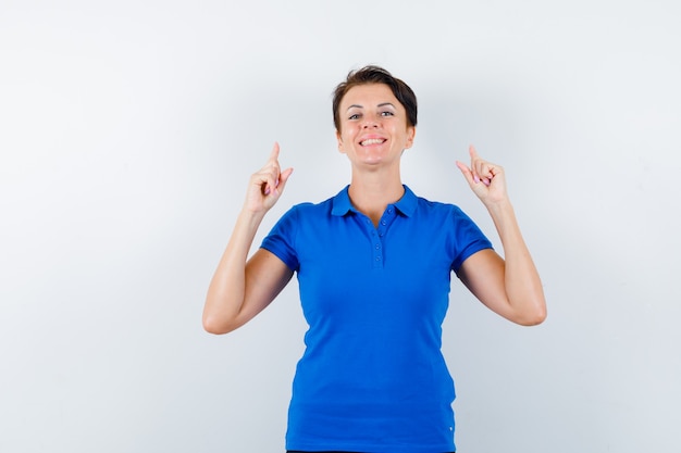 Mulher madura apontando para cima em t-shirt azul e parecendo confiante. vista frontal.