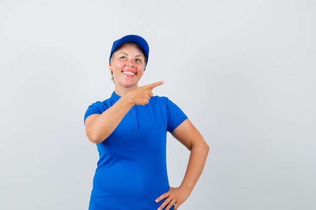 Mulher madura, apontando para o lado em t-shirt azul e olhando alegre.