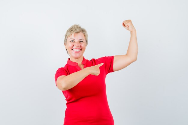 Mulher madura apontando para os músculos do braço em uma camiseta vermelha e parecendo confiante