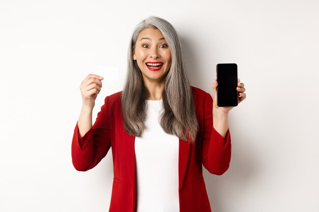 Mulher madura asiática alegre mostrando a tela do smartphone em branco e cartão de crédito, conceito de e-commerce.