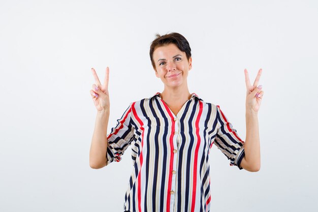 Mulher madura com camisa listrada, mostrando o gesto de paz e olhando feliz, vista frontal.